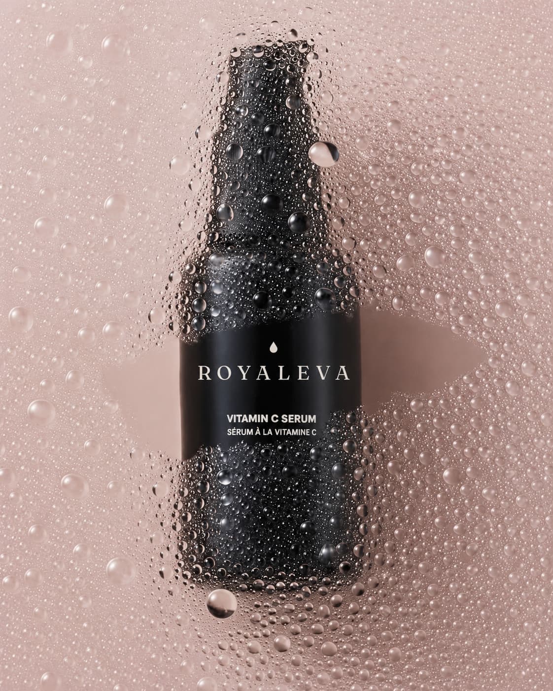 A black Royaleva vitamin C serum bottle seen through a veil of water droplets on a pale pink ground.