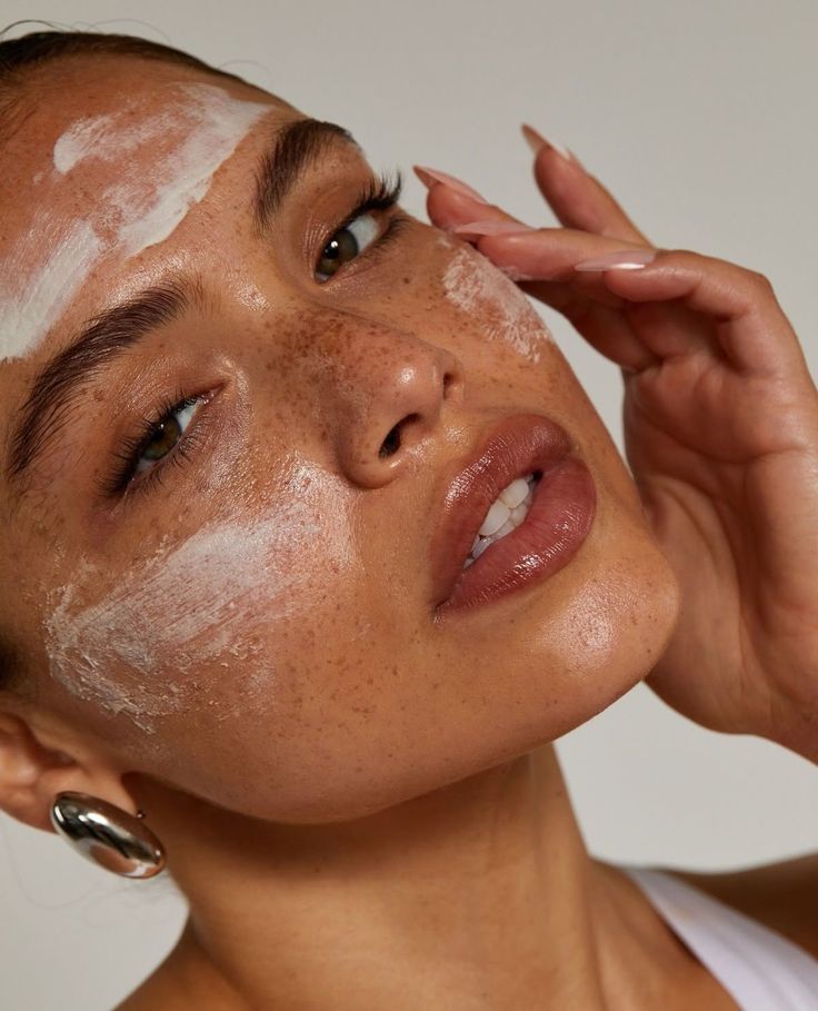 A face in three quarters with cream spread in patches across the forehead and cheek, a fingertip pressing it in.