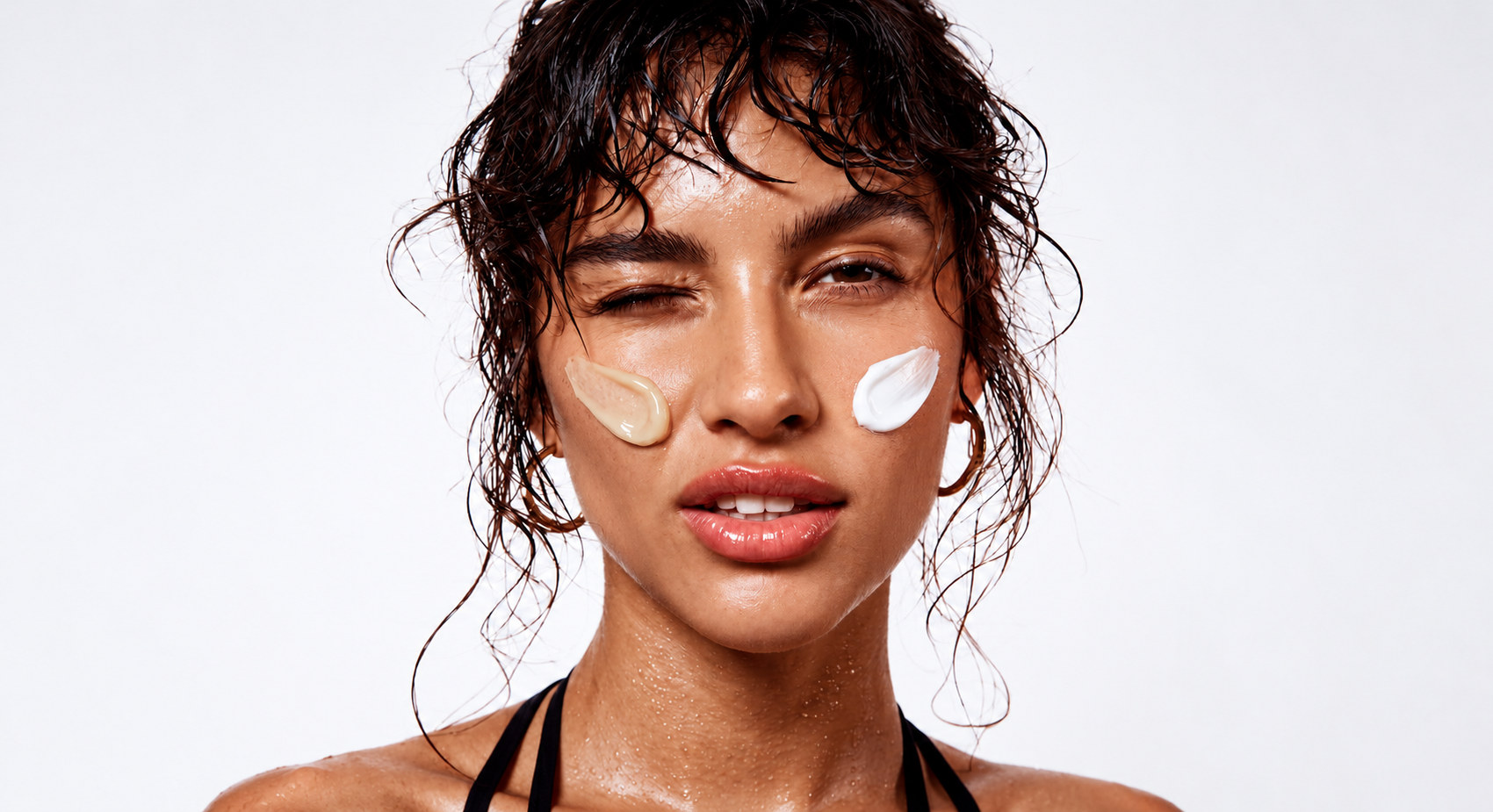 Portrait with wet hair, two cream swatches on the cheeks, on a pale ground.